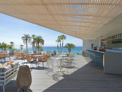 Bar sulla spiaggia aperta con tavoli, sedie e vista su palme e mare sotto il sole.