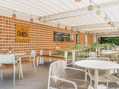 Terrazza moderna e soleggiata con tavoli e sedie bianchi di fronte a un muro di mattoni ben illuminato.