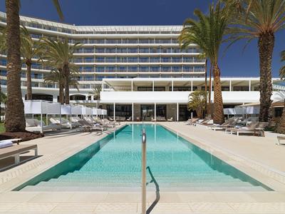 Hotel moderno con una piscina lunga, circondato da palme e sdraio sotto un cielo azzurro.