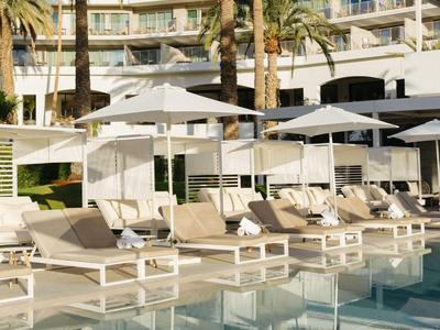 Piscina dell'hotel con lettini, ombrelloni e palme davanti a un edificio pluripiano.