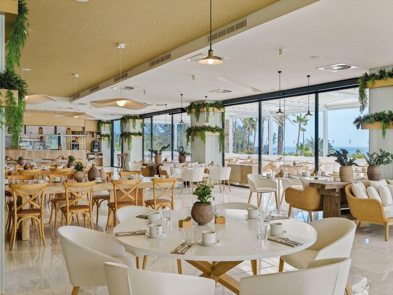 Ristorante luminoso e moderno con grandi finestre, piante e tavoli bianchi con sedie.