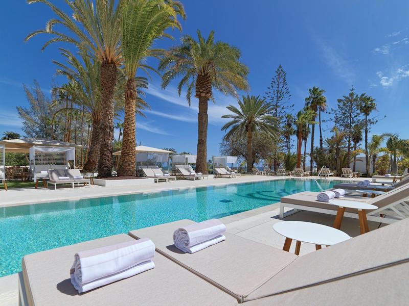 Area piscina moderna con palme, lettini e cielo azzurro in un resort alberghiero.