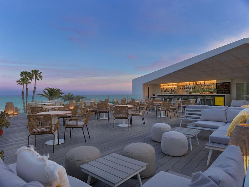 Terrazza moderna dell'hotel con mobili lounge e bar, vista sul mare e palme al tramonto.
