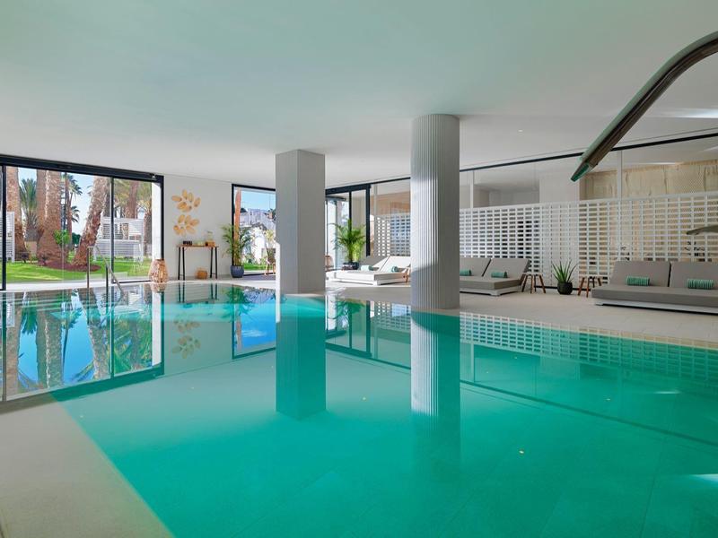 Piscina interna con colonne, lettini e grandi finestre con vista su palme.