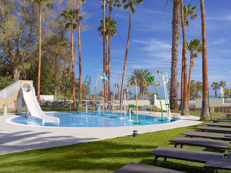 Piscina dell'hotel con scivoli d'acqua, lettini e alti palme sotto un cielo azzurro.