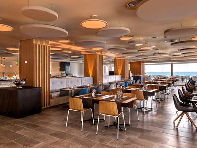 Moderner, heller Restaurantraum mit Holz, runden Deckenleuchten und großen Fenstern am Meer.