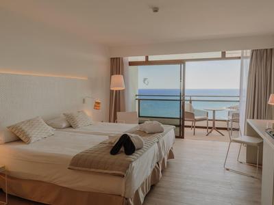 Helles Hotelzimmer mit zwei Betten, Balkon und Meerblick, weiße Wände, Holzboden und moderne Möbel.