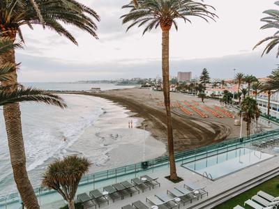Strand mit Palmen, Wellen und einem Schwimmbecken neben einem Hotel am Meer bei bewölktem Himmel.