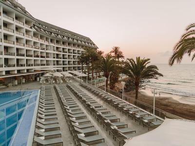 Hotel mit geschwungenem Bau, Pool, vielen Liegen und Palmen am Strand bei Sonnenuntergang.