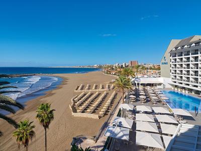 Hotel con piscina y sombrillas en la playa con vistas al mar y cielo azul.