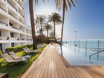 Piscina moderna de hotel con terraza de madera, tumbonas y palmeras con vista al mar bajo cielo despejado.