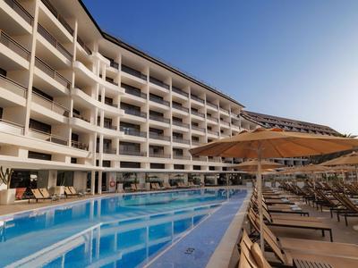 Hotel moderno con gran piscina al aire libre y tumbonas bajo sombrillas en día despejado.