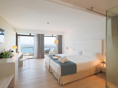 Habitación de hotel luminosa con cama doble, escritorio, sillas y vista al mar a través de grandes ventanas.
