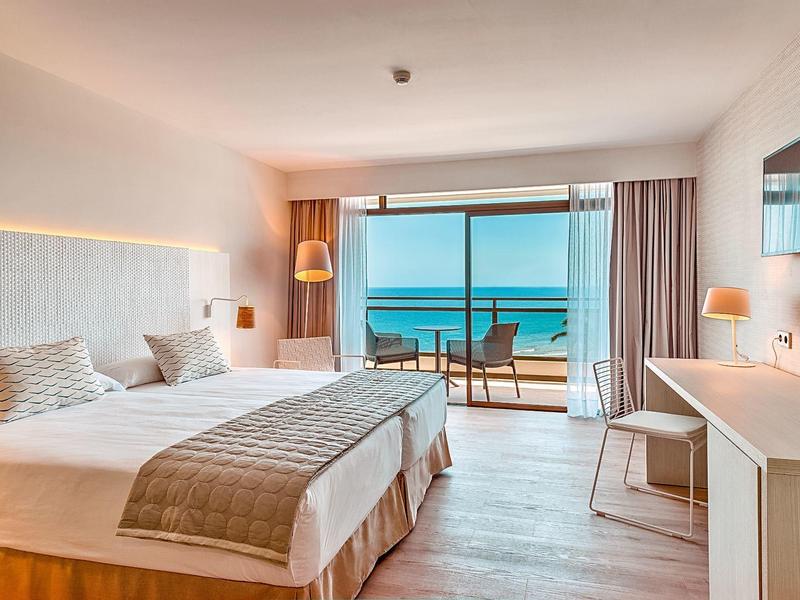 Bright hotel room with twin beds, balcony overlooking the ocean, and modern decor.