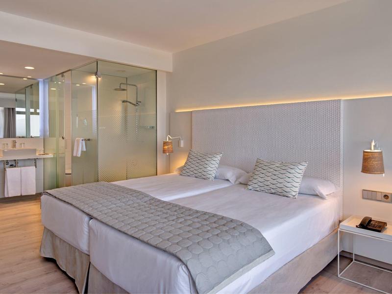 Modern hotel room with a large bed, glass shower, and minimalist decor in soft neutral tones.