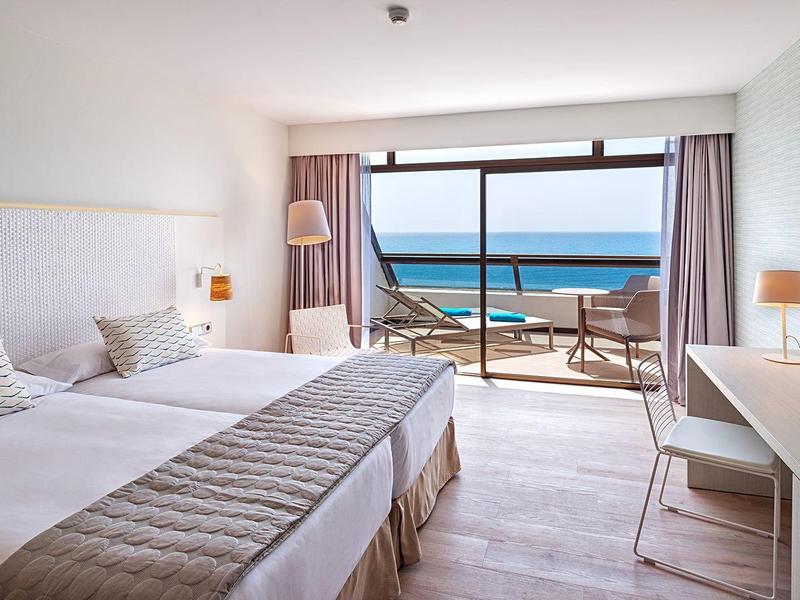 Hotel room with twin beds, balcony overlooks ocean and sandy beach under clear sky.