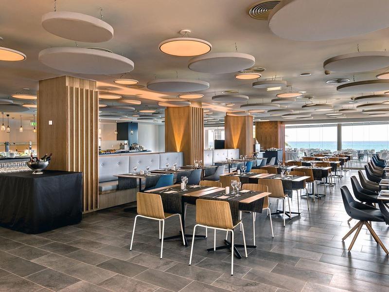 Spacious hotel dining area with modern decor, wooden tables, cushioned chairs, and large windows.