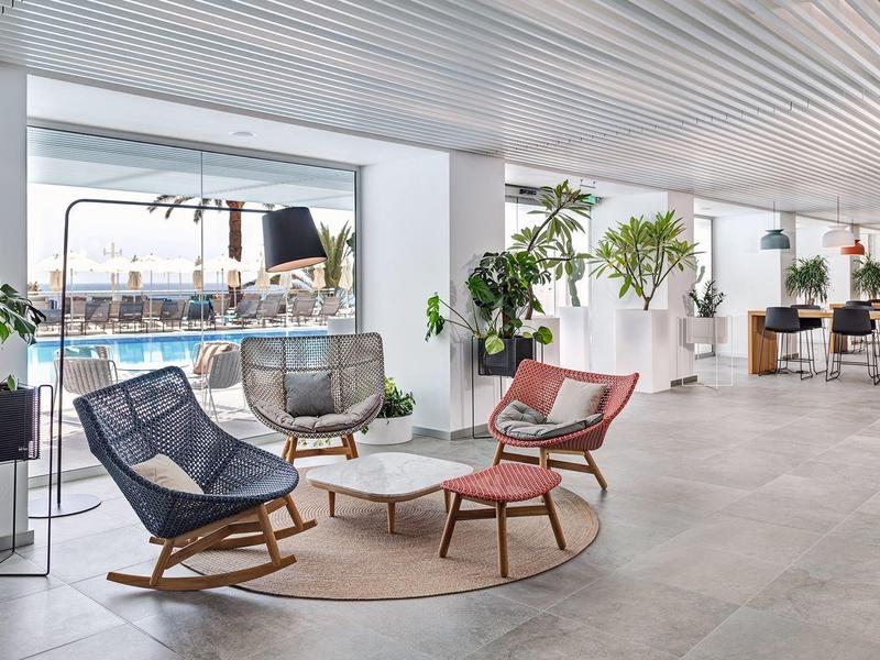 Modern hotel lobby with lounge chairs, plants, and a view of the beach and ocean.