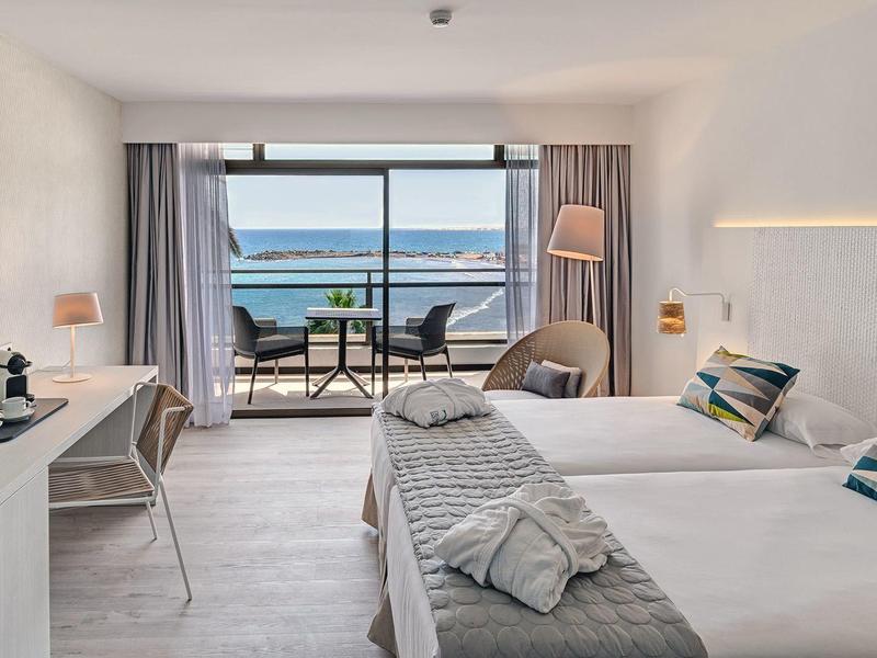 Hotel room with two beds, balcony overlooking sea and hills, and modern decor.