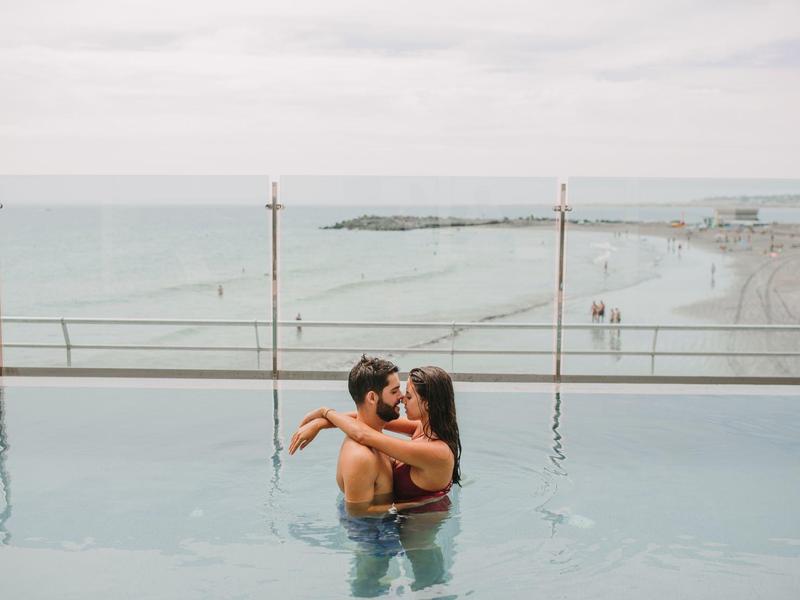 Couple embracing in an infinity pool overlooking a beach and ocean under cloudy sky.