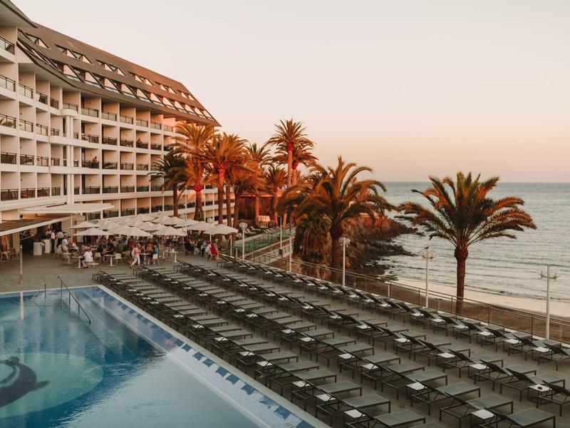 Resort with rows of poolside lounge chairs, palm trees, and ocean views during sunset.