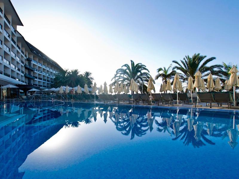 Outdoor pool with umbrellas and palm trees next to a hotel building under clear sky.