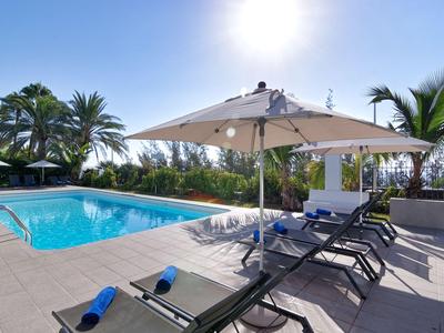 Słoneczny obszar przy basenie z leżakami, parasolem i palmami w hotelowym ogrodzie.