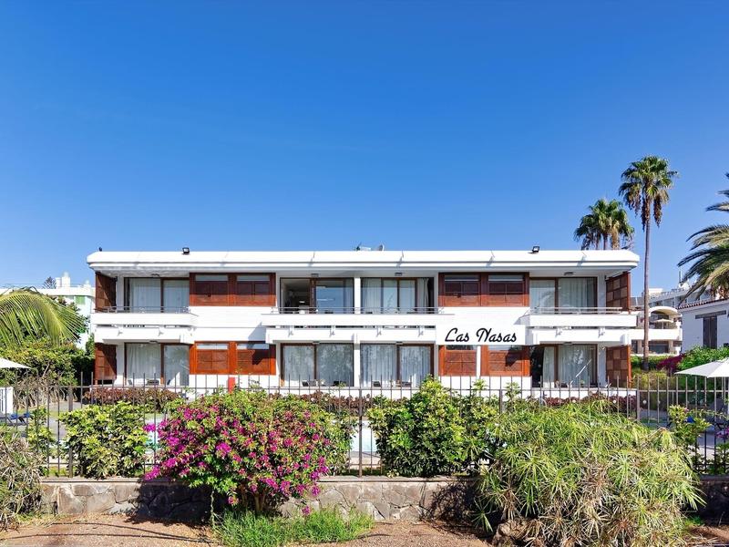 Modern hotel building with white walls, balconies, and palm trees under a clear blue sky.
