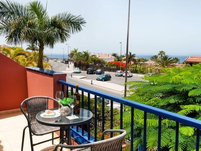 Balkon z stołem i krzesłami z widokiem na ulicę, palmy i morze w tle.