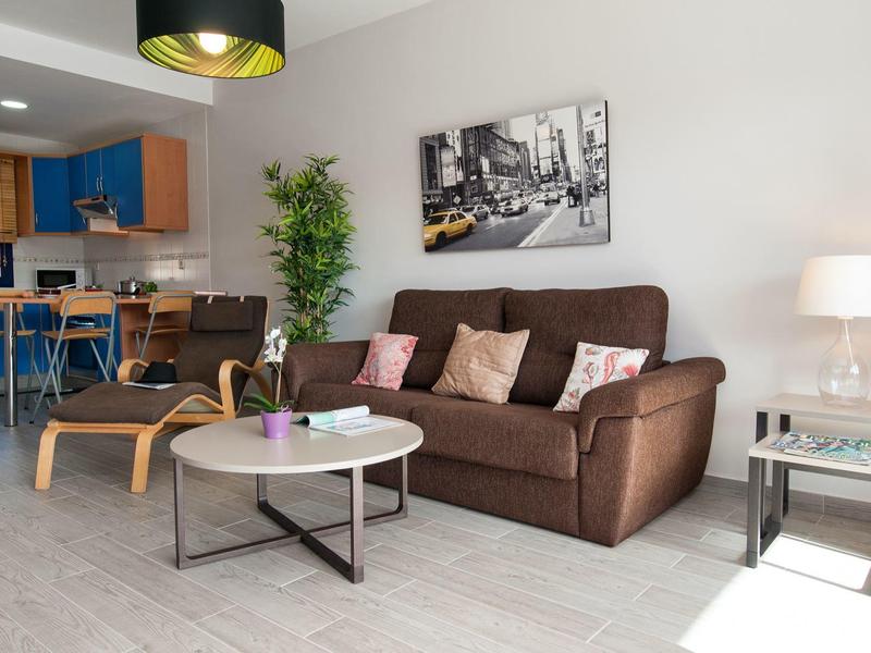 Salon confortable avec canapé marron, fauteuil, table basse et coin cuisine moderne.