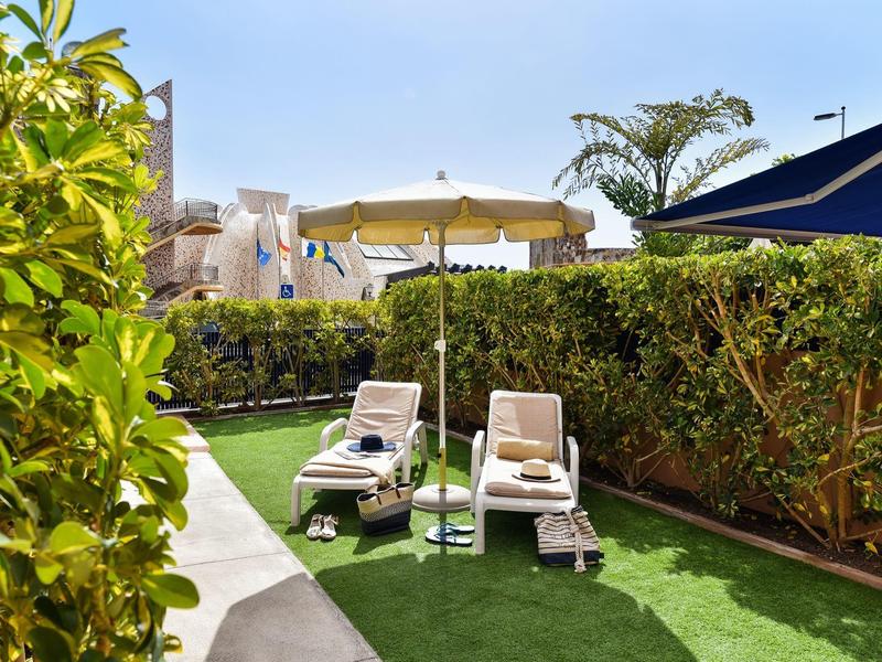 Deux chaises longues sous un parasol dans un jardin d'hôtel avec herbe verte et haie.