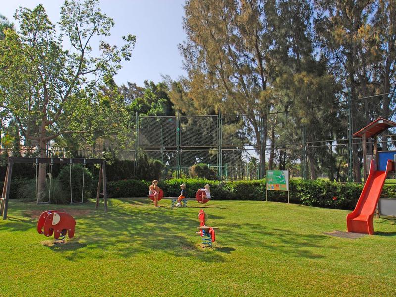 Área de juegos infantiles con columpios, tobogán y figuras para jugar en un jardín verde.