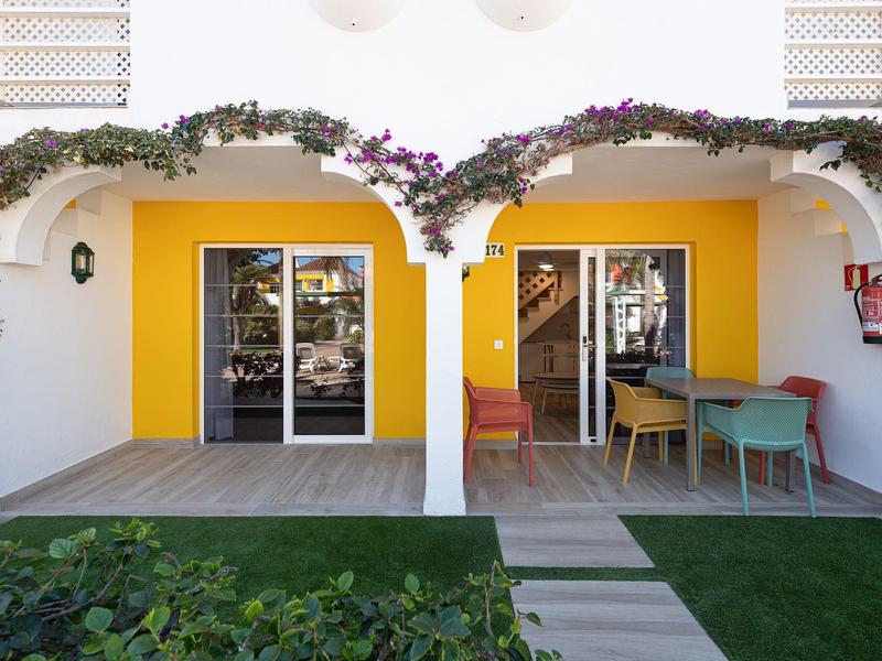 Área exterior de un hotel con paredes amarillas, dos puertas y balcón amueblado con plantas.