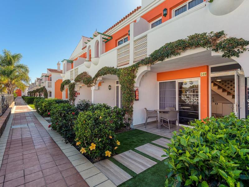 Casas en fila con terrazas, jardín verde y toldos bajo cielo despejado junto a una acera.