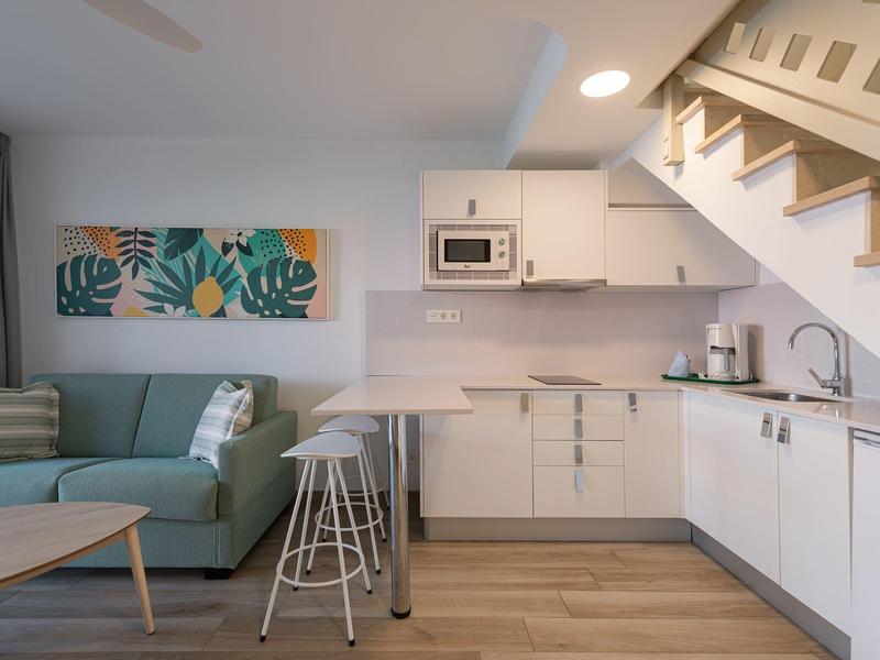 Cocina luminosa con muebles blancos, taburetes altos y un sofá verde en el área de estar bajo la escalera.