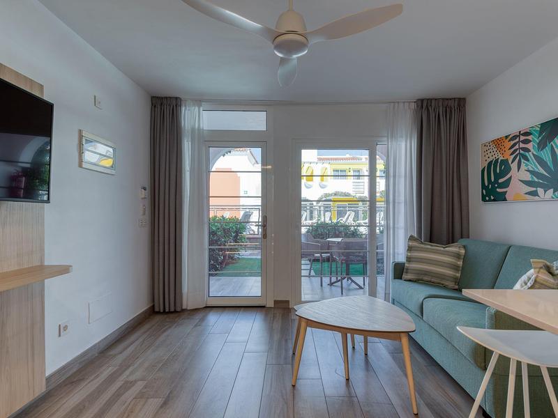 Sala de estar luminosa con sofá, mesa de centro y televisor, vista a terraza con jardín.