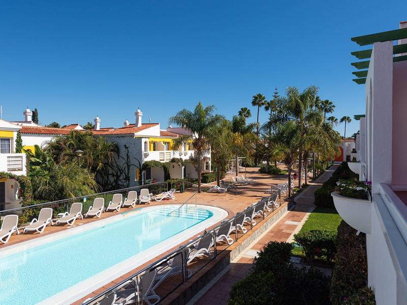 Piscina al aire libre rodeada de tumbonas junto a edificios de hotel y palmeras bajo un cielo azul.