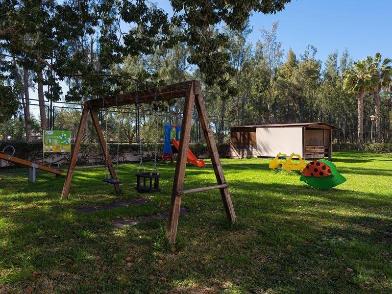 Un parque infantil con columpios en un césped verde junto a un edificio de madera, rodeado de árboles.