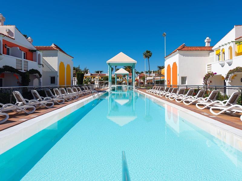 Piscina moderna de hotel con tumbonas, rodeada de edificios blancos y amarillos bajo un cielo azul claro.