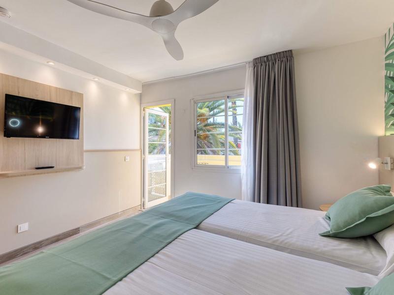 Habitación de hotel moderna con dos camas, acentos verdes y ventilador de techo a la luz del día.
