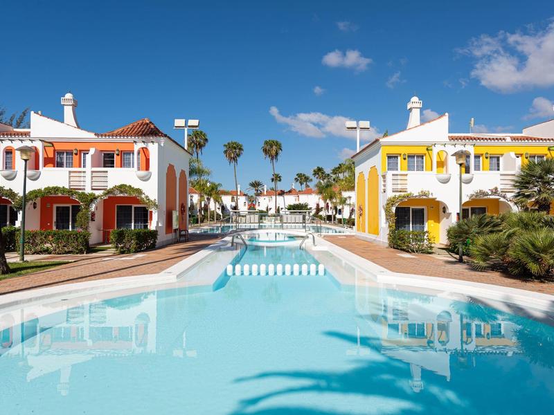 Dos casas coloridas junto a una piscina larga bajo un cielo azul con palmeras.