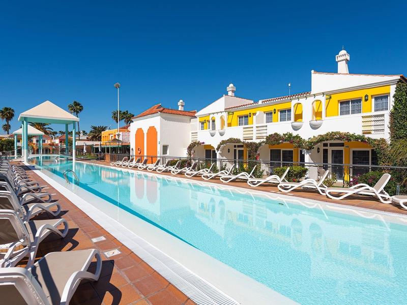 Una piscina al aire libre con tumbonas y un hotel de varios pisos al fondo bajo un cielo soleado.