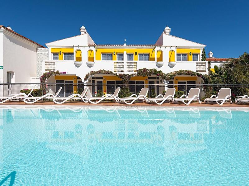 Hotel con acentos amarillos claros, piscina y filas de tumbonas blancas bajo cielo despejado.