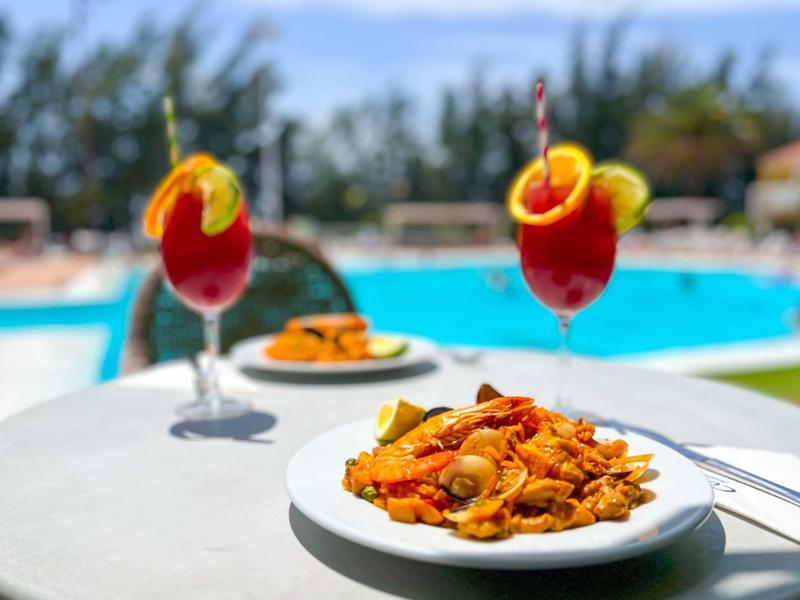 Plato de pasta con mariscos sobre una mesa blanca junto a una piscina con bebidas y sombrillas rojas.