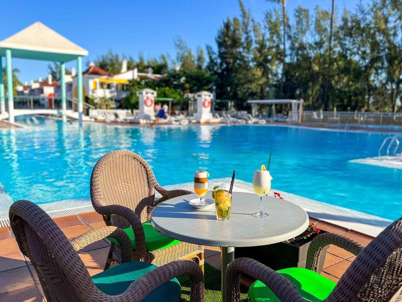 Piscina tropical de hotel con mesa redonda y cuatro sillas, bebidas sobre la mesa, día soleado.
