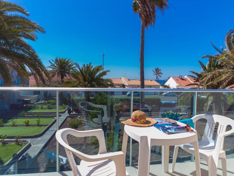 Balcone con sedie bianche e tavolo con cappello e riviste, vista su palme e case.