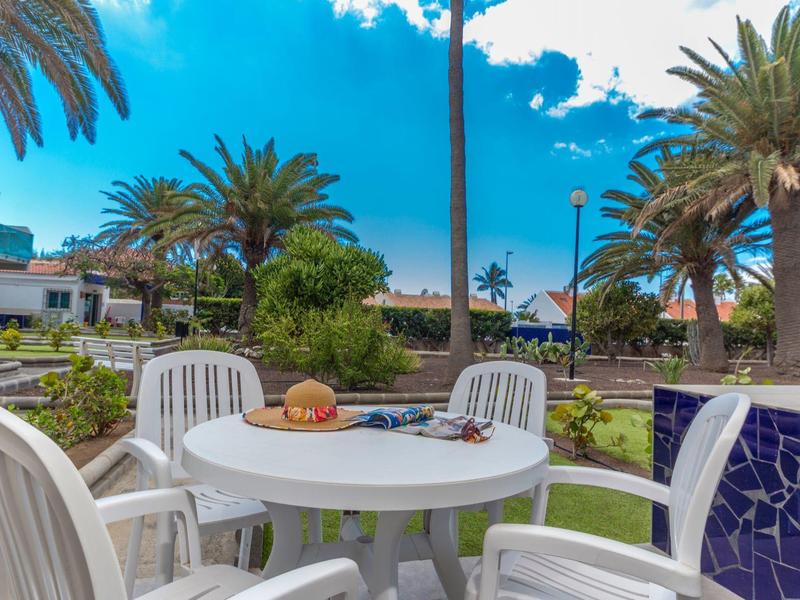 Tavolo da patio con sedie, decorato con pane e un giornale, circondato da palme.