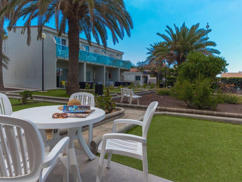 Un'area esterna con sedie di plastica bianche e un tavolo rotondo accanto a un prato verde e palme.