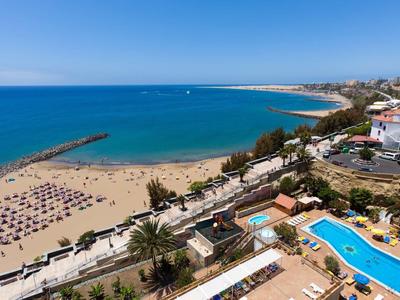 Blick auf einen Sandstrand mit Sonnenliegen, blauen Meer und Poolbereich in einer Küstenstadt.