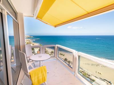 Balkon mit Glaskante, gelbem Sonnenschutz und kleinem Tisch, Blick auf Meer und Strand.